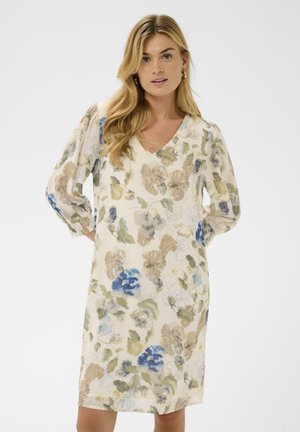 Kvinde med langt blondt hår iført en knælængde cremet kjole med blå, grønne og beige blomsterprint mod en ensfarvet hvid baggrund.