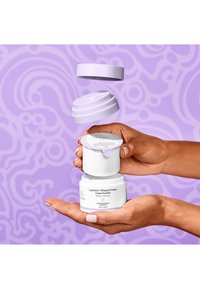 White jar with rounded design and a light purple lid, held in a hand. Label features product name and key details, against a soft purple background.