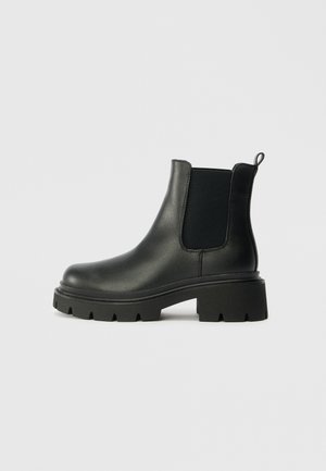Bottines Chelsea en cuir noir avec des panneaux élastiques sur les côtés, une finition texturée et une semelle en caoutchouc épaisse. Comprend une languette pour un enfilage facile.