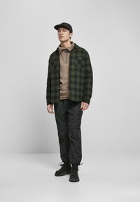 Czarna i zielona koszula w kratę z guziczkami, nałożona na beżowy sweter, zestawiona z czarnymi zwężającymi się spodniami oraz czarnymi butami na sznurowadła.