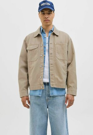 Beige katoenen jas met twee borstzakken, knoopsluiting en brede mouwen, gelaagd over een denim overhemd, gecombineerd met lichtblauwe jeans.