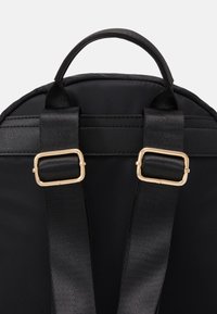 Sac à dos noir avec tissu lisse, pièces en métal doré, bretelles rembourrées et réglables, et poignée supérieure arrondie pour un transport facile.