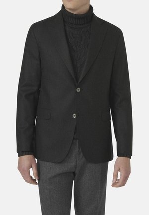 Schwarzer maßgeschneiderter Blazer aus einem glatten Stoff, mit zwei Knöpfen, Reverskragen und Seitentaschen, über einem dunklen Rollkragenpullover getragen.