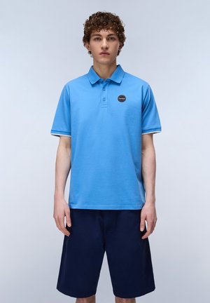 Jonge man met krullend haar, gekleed in een lichtblauw poloshirt met een klein logo op de borst en donkerblauwe shorts tot op de knie, staand tegen een effen achtergrond.