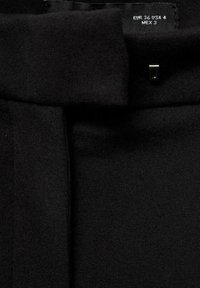 Pantalon en tissu noir avec une texture lisse, des coutures visibles et un petit élément métallique, avec une étiquette d'entretien indiquant la taille.