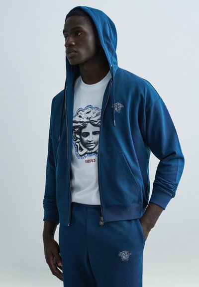 Blue zip-up hoodie with drawstrings, featuring a Medusa logo on the chest. Paired with a white T-shirt displaying a graphic print and logo.