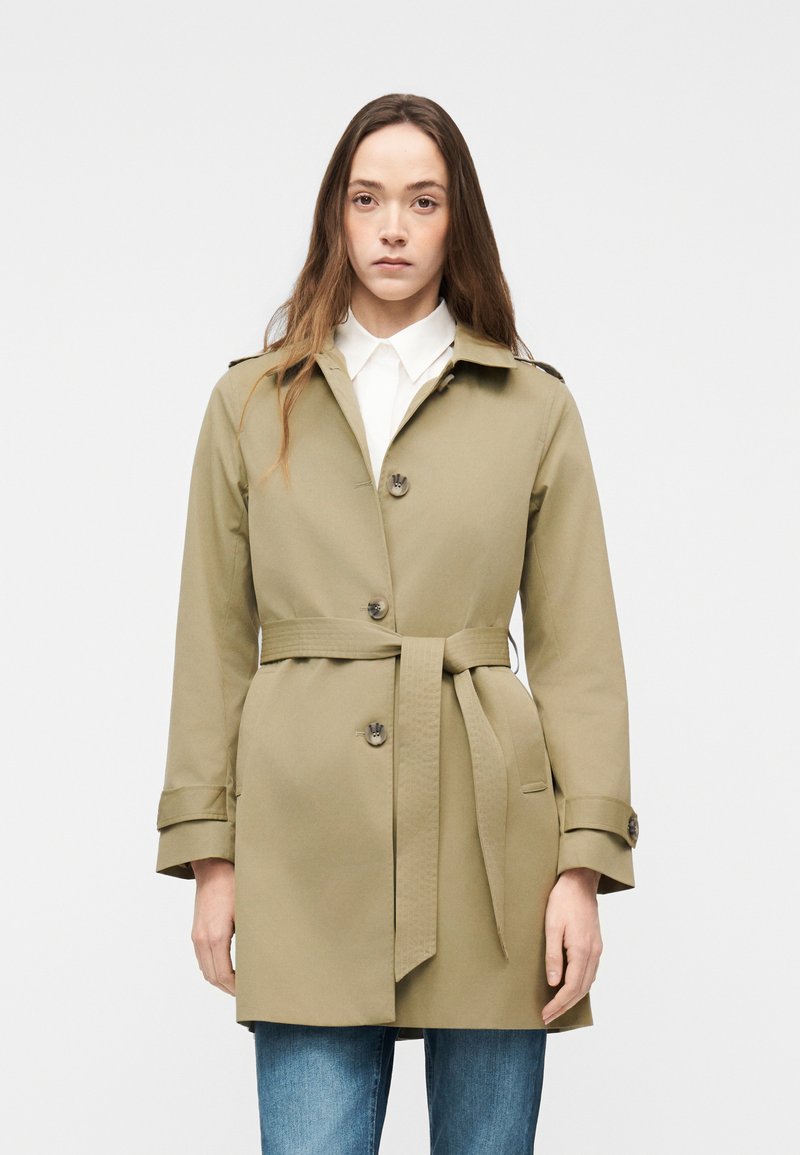 Jeune femme portant un trench-coat beige ceinturé par-dessus une chemise blanche et un jean bleu, debout devant un fond clair uni.