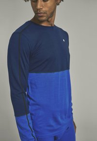 Long-sleeve shirt in dark and light blue, featuring a color block design with black stitching. Fabric appears smooth with slight texture.