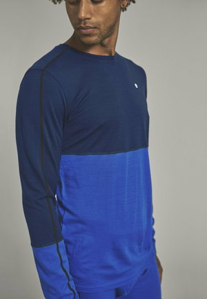 Long-sleeve shirt in dark and light blue, featuring a color block design with black stitching. Fabric appears smooth with slight texture.