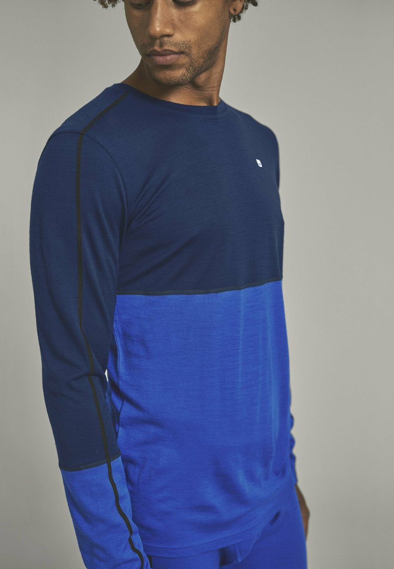 Long-sleeve shirt in dark and light blue, featuring a color block design with black stitching. Fabric appears smooth with slight texture.