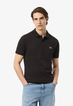 Giovane con capelli castani mossi, indossa una polo nera a maniche corte con un piccolo logo di coccodrillo verde e jeans azzurri chiari, mani nelle tasche.