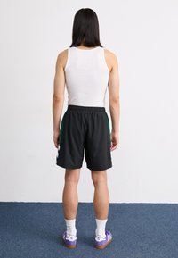 Svarte sportshorts med grønne side-striper, kombinert med en hvit tank topp og lilla sko. Bakgrunnen har en vanlig vegg og blå teppe.