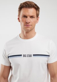 Balta medvilnės marškinėliai su juodu tekstu "BIG STAR", virš ir žemiau teksto yra tamsiai mėlyni ir šviesiai mėlyni horizontalaus rašto dryžiai.