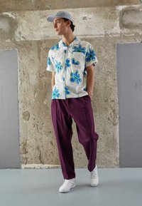 Kortärmad skjorta i vitt med blå och gröna blommönster; kombineras med löst sittande lila byxor och vita sneakers.