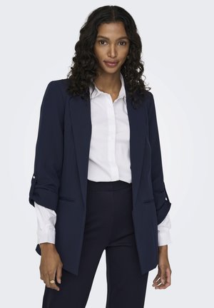 Blazer navy en tejido ligero, con un corte relajado, solapas puntiagudas y mangas enrolladas con sutiles botones. Camisa blanca de botones debajo.