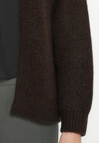 Brun strikket cardigan med en teksturert finish, som har ribbestrikkede mansjetter og en avslappet passform, kombinert med en svart topp og olivengrønne bukser.