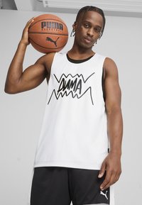 Jersey de baloncesto blanco sin mangas con ribete negro y un atrevido logo negro de "PUMA" junto con un patrón en zigzag. El modelo sostiene un balón de baloncesto sobre su hombro.