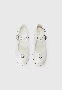 White Mary Jane shoes featuring a rounded toe, buckle strap, metallic accents, and black studs along the sole for texture and design detail.