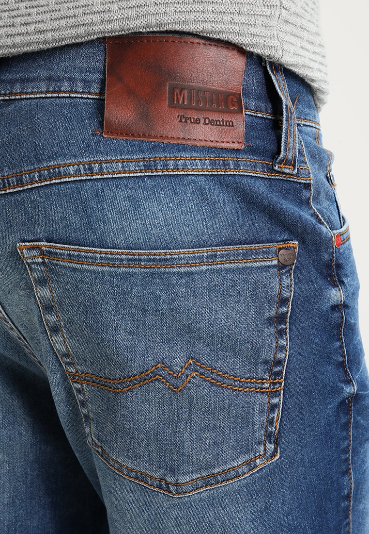 Blaue Jeans aus Denim mit orangefarbenen Nähten, einem braunen Lederpatch mit der Aufschrift "MUSTANG True Denim" und einem markanten Taschendesign.