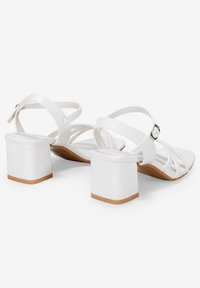 Sandalias de tacón blancas con tacón cuadrado, tiras delgadas y una correa en el tobillo con hebilla, con un acabado suave y suela de color marrón claro.
