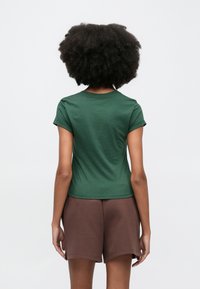 Πράσινο βαμβακερό κοντομάνικο μπλουζάκι με στρογγυλή λαιμόκοψη και εφαρμοστή σχεδίαση, συνδυασμένο με καφέ αθλητικές βερμούδες, από πίσω όψη.