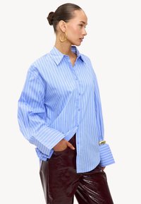 Lichtblauwe overhemdbloes met witte verticale strepen, met een klassieke kraag en oversized manchetten, gecombineerd met glanzende bordeauxrode pantalons.