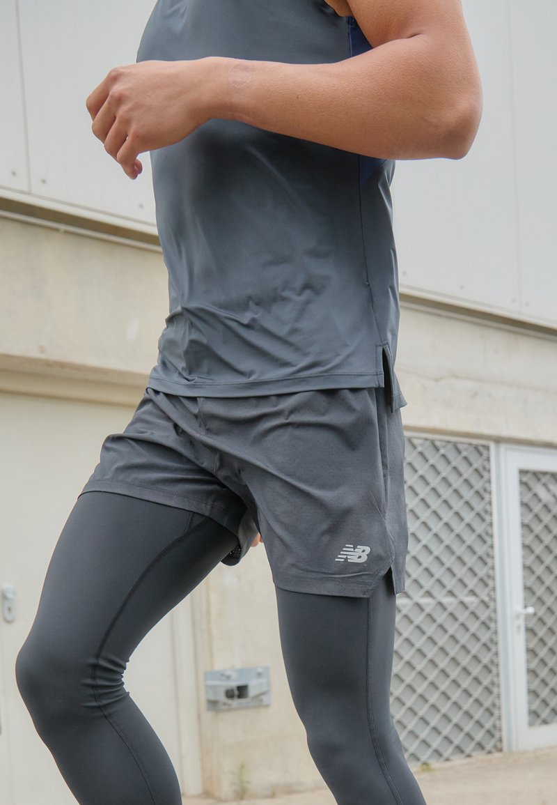 Débardeur de sport gris et shorts, associés à des leggings noirs. Le matériau semble léger et respirant ; comporte le logo New Balance.