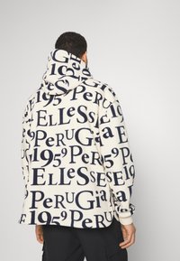 La sudadera de forro polar presenta un fondo crema con texto en azul oscuro, incluyendo logotipos y números, capucha y puños acanalados.
