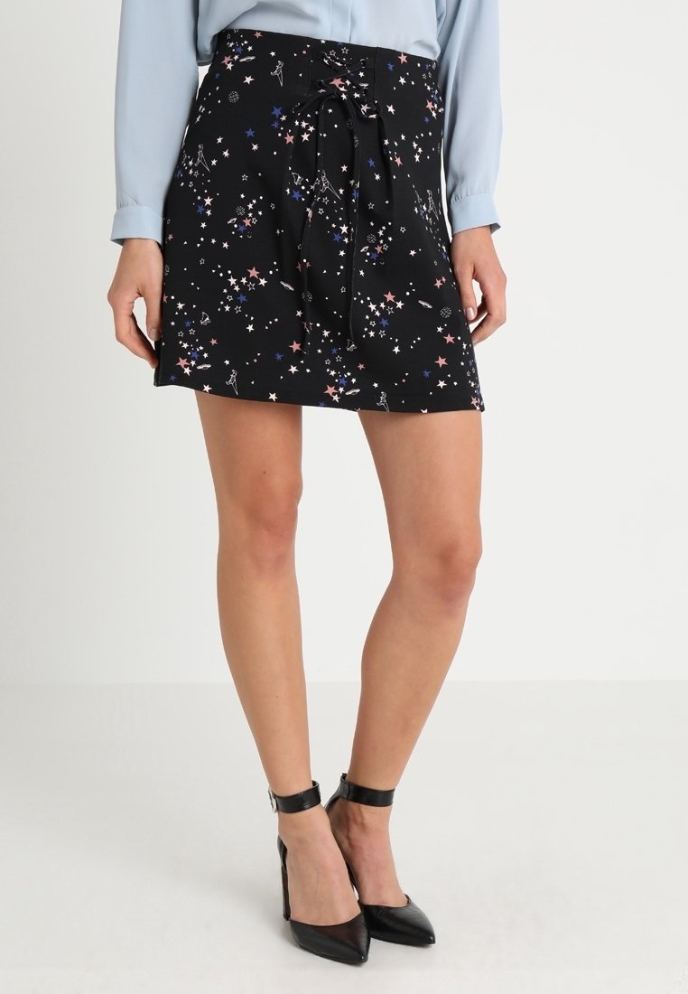 Mini-jupe noire avec motifs colorés d'étoiles et de constellations, ceinture nouée à la taille, portée avec un chemisier bleu et des talons noirs à bride cheville.