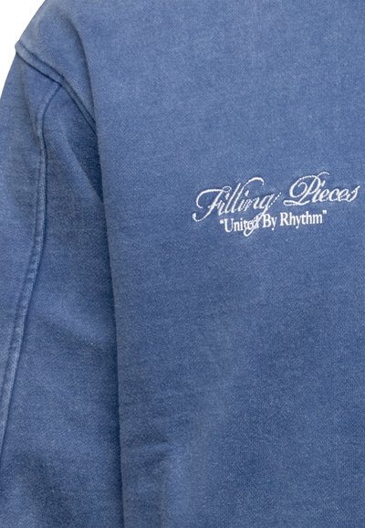Blue sweatshirt made from soft fabric, featuring white embroidered logo "Filling Pieces" and tagline "United By Rhythm" on the chest.