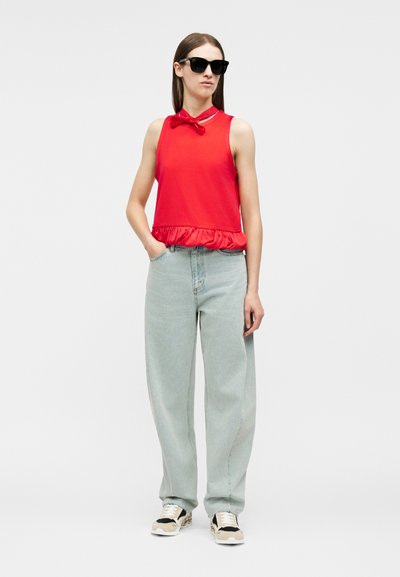 Γυναίκα που φοράει μαύρα γυαλιά ηλίου, κόκκινο αμάνικο τοπ με φιόγκο στο λαιμό, ανοιχτό μπλε άνετο τζιν και αθλητικά παπούτσια, στέκεται με το ένα χέρι στην τσέπη.