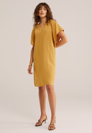 Nő áll, rajta egy mustársárga térdig érő ruha rövid fodros ujjakkal, és nyitott orrú magas sarkú szandál.