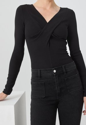 Vrouw draagt een nauwsluitende zwarte top met lange mouwen en een gedraaide V-hals en hooggetailleerde zwarte jeans, leunend tegen een witte blok.