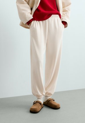 Personne portant un pantalon de jogging beige, des chaussettes blanches, des chaussures à enfiler marron avec boucle, un pull rouge et une veste beige, les mains dans les poches.