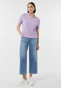 Lila Kurzarm-Strick-Polohemd mit V-Ausschnitt, kombiniert mit hellblauen weiten Jeans und weißen Turnschuhen, vor einem neutralen Hintergrund stehend.