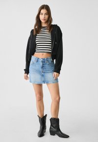 Cropped hoodie nera, top a righe bianche e nere, mini gonna di denim blu e stivali da cowboy neri con un lieve tacco. Presentazione di un outfit casual.