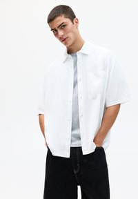 Chemise blanche à manches courtes avec une poche avant, superposée sur un t-shirt gris. Pantalon en denim noir avec des coutures blanches contrastées.