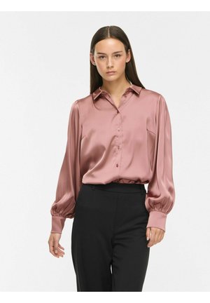 Frau mit glatten dunklen Haaren, die eine langärmelige glänzende rosa Satinbluse und schwarze hoch taillierte Hose trägt, steht vor einem schlichten Hintergrund.