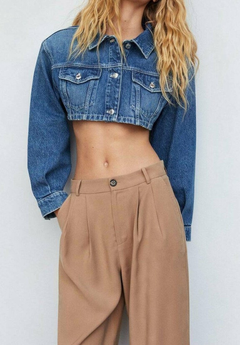 Femme portant une veste en jean bleue courte et un pantalon beige taille haute, mains dans les poches, debout devant un fond clair uni.