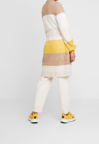 Długa do kolan, paskowana kardigan w kolorach beżowym, białym i żółtym, o miękkiej fakturze. Sparowana z kremowymi spodniami i żółtymi trampkami.