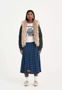 La modella indossa una giacca bomber verde scuro con un colletto beige soffice, una t-shirt bianca con grafica e una gonna midi a quadri blu con un motivo accentuato.