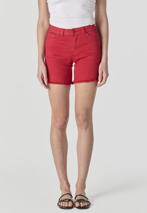 Mujer con pantalones cortos de mezclilla rojos con bordes deshilachados, una camiseta sin mangas blanca y sandalias negras planas, de pie contra un fondo liso.