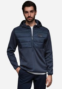 Marinblå huva jacka med en kviltad övre del och texturerad underdel, med en framdragkedja och en liten logotyp-patch.