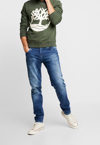 Felpa verde oliva con logo bianco a forma di albero, abbinata a jeans in denim blu. Il modello indossa sneaker bianche e si trova in piedi con una mano in tasca.