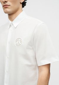 Νεαρός άνδρας που φοράει ένα λευκό κοντομάνικο πουκάμισο με κουμπιά και στρογγυλό λογότυπο της Versace Jeans Couture στο στήθος.