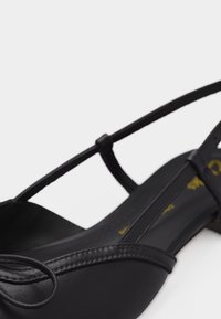 Μαύρο δερμάτινο επίπεδο παπούτσι με λεπτό λουράκι και μυτερή μύτη, που διαθέτει ένα λεπτεπίλεπτο μικρό φιόγκο στο μπροστινό μέρος.