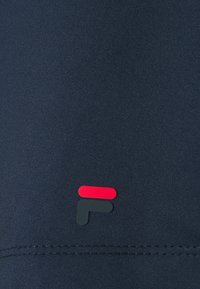 Tejido azul marino con una textura suave que presenta un pequeño logo en rojo y verde oscuro en la parte inferior. Detalle de costura visible.