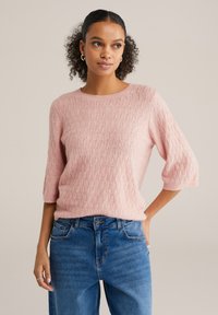 Pulover tricotat roz deschis cu un model texturat în formă de diamant și mâneci scurte, asortat cu blugi din denim albastru cu talie înaltă.