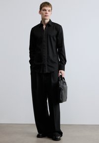 Fekete hosszú ujjú ing texturált gombolósávval, hozzá fekete bő szárú nadrág és fekete cipő. A modell szürke kézitáskát tart a kezében.