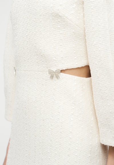 Λευκό φόρεμα με υφή και σκίσιμο στη μέση, διακοσμημένο με μια μικρή ασημένια φιόγκου.